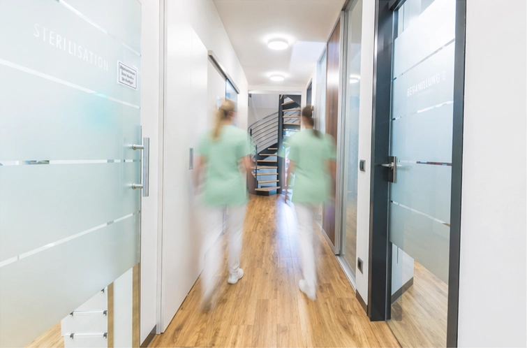 Zwei Mitarbeitende in mintgrüner Praxiskleidung gehen durch den hellen Flur von DEIN DENTAL Stammham.